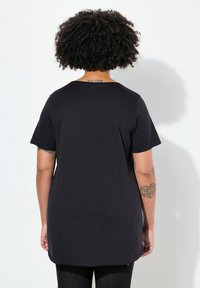 Camiseta negra de manga corta, hecha de algodón suave, con un ajuste relajado y un dobladillo redondeado, con tatuajes visibles en los brazos.