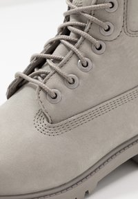 Stivale alla caviglia in suede grigio con occhielli metallici rotondi, lacci grigi, dettagli di cuciture laterali e suola in gomma testurizzata.