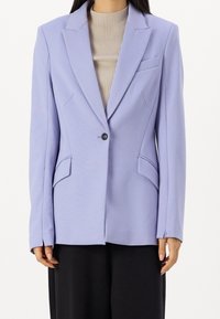 Donna che indossa un blazer a un bottone di colore lilla chiaro sopra un top rigato beige e pantaloni neri, in piedi contro uno sfondo bianco semplice.