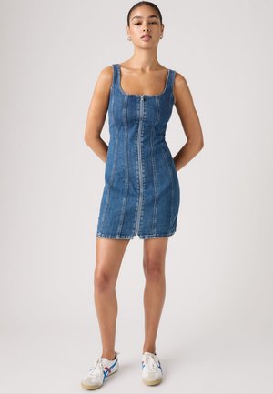 Donna in piedi con le mani dietro la schiena, indossa un vestito aderente in denim blu senza maniche con cerniera frontale e sneaker bianche con strisce blu e rosse.