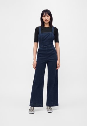 Junge Frau steht und trägt dunkelblaue, weit geschnittene Denim-Latzhosen über einem schwarzen Kurzarmtop und schwarz-weiße karierte Slip-On-Schuhe.