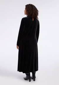 Černé sametové šaty s dlouhými rukávy, bílým puntíkovým vzorem, véčkovým výstřihem na zádech a splývavou spodní částí, doplněné černými kozačkami ke kolenům.