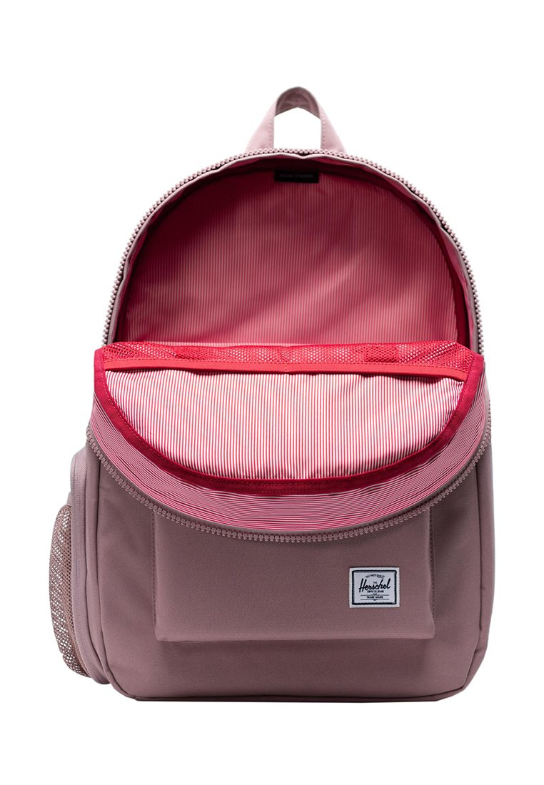 Rucksack aus schlichter rosafarbener Stoff mit Reißverschluss. Das Innere hat ein gestreiftes rotes Futter und eine Netztasche. Auf der Vorderseite befindet sich ein kleines Logo-Patch.