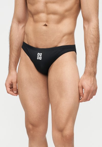 Ανδρικός κορμός με μαύρο μαγιό μπριφ με λευκό λογότυπο "D2" μπροστά, στέκεται μπροστά σε απλό ανοιχτό φόντο.