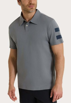 Uomo che indossa una polo grigia a maniche corte con due strisce scure sulla manica sinistra, abbinata a pantaloni neri, torso visibile.