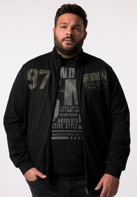 Black zip-up jacket featuring a stand-up collar and printed text in gray. Worn over a black graphic t-shirt with white text and patterns.