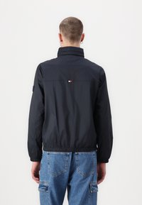 Jachetă tip windbreaker de culoare albastru marin, cu guler înalt, manșete elastice și talie ajustată. Dispune de un mic logo roșu, alb și albastru pe spate.