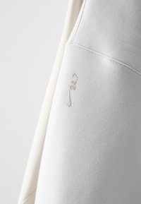 Sweatshirt Nike em tecido bege claro. Apresenta uma textura suave, logotipo em cinza claro e um detalhe de costura. Número de modelo "24/7" na manga.
