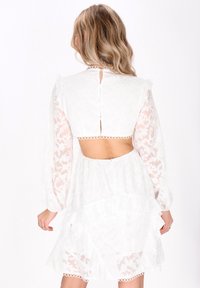 Rochie albă cu mâneci lungi transparente și spate deschis. Fustă cu volane stratificate și textură din dantelă florală. Închidere cu nasturi la gât.