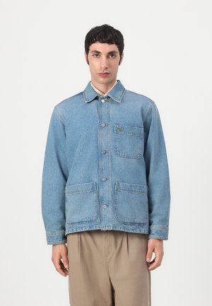 Jeune homme portant une veste en jean bleu clair boutonnée avec trois poches devant et un pantalon beige sur un fond uni.