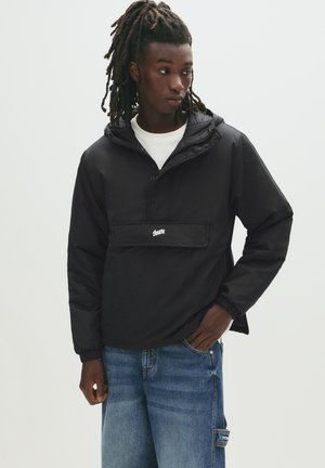 Schwarze Kapuzenanorakjacke mit elastischen Bündchen und einer Fronttasche. Hergestellt aus leichtem Material, mit einem Logo unterhalb der Tasche.