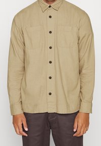 Camicia a maniche lunghe marrone chiaro con abbottonatura frontale, due tasche sul petto e colletto. Il tessuto sembra texturizzato con una vestibilità comoda.