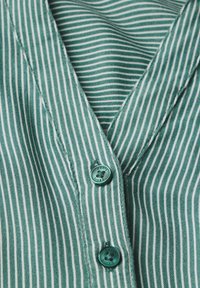 Tessuto a strisce verdi con design a scollo a V, presente due bottoni turchesi e una texture sottile. Le strisce sono distanziate uniformemente e verticali.