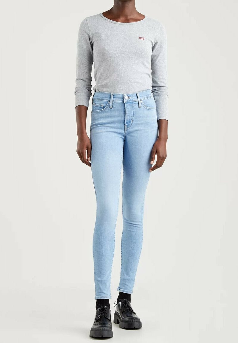 Personne portant un jean skinny bleu clair, un t-shirt gris à manches longues, des chaussettes noires et des chaussures noires épaisses sur fond uni.