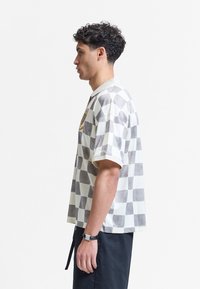 Jeune homme aux cheveux bouclés portant une chemise à manches courtes à carreaux, un pantalon noir et une montre-bracelet, debout de profil sur fond blanc.