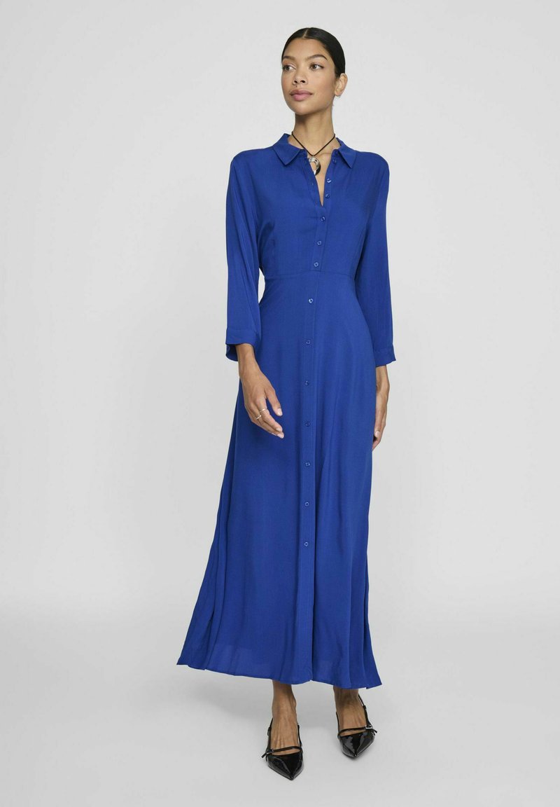 Mėlyna ilga rankovė suknelė, pagaminta iš lygaus audinio, su mygtukų užsegimu priekyje, apykakle ir šoniniu įpjovimu, derinama su juodais smailiais bateliais.