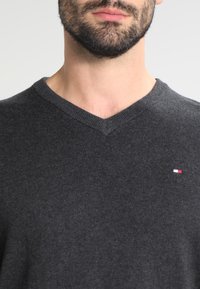 Tummanharmaa V-kaula-aukkoinen neule, jossa on neulottu pinta ja pieni logo punaisilla, valkoisilla ja sinisillä yksityiskohdilla vasemmassa rinnassa. Yksinkertainen, vartalonmyötäinen malli.