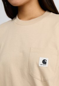 T-shirt en coton beige avec un col rond, dotée d'une poche sur la poitrine gauche avec une étiquette blanche affichant un logo noir. Texture douce, design décontracté.