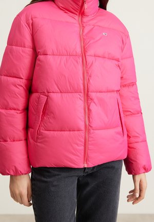 Person som bär en klar rosa pufferjacka med frontdragkedja och fickor, ihop med svarta byxor, korta vid midjan och armarna synliga.