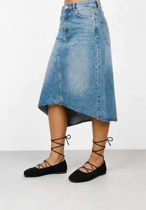 Falda midi asimétrica de denim con dobladillo sin rematar combinada con bailarinas negras con cordones en una persona de pie.