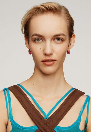 Modelo vistiendo una blusa turquesa con tirantes cruzados marrones y pendientes de aro rosas. Tiene el cabello corto y de color claro, con un maquillaje natural.