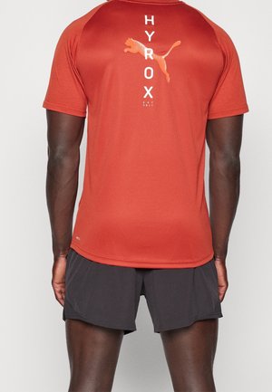 Mężczyzna noszący pomarańczowy, przypominający spalony, krótki top Puma Hyrox i czarne sportowe spodenki, widziany od tyłu na jednolitym tle.