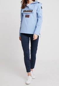 Jasnoniebieska wiatrówka z przednią kieszenią i naszywką z flagą Norwegii, w zestawieniu z ciemnoniebieskimi spodniami typu slim-fit i białymi trampkami.