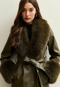 Oliwkowy skórzany płaszcz z dużym, luksusowym kołnierzem i mankietami z sztucznego futra. Posiada szarfę w talii oraz wyraźnie zarysowaną sylwetkę.