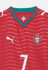 Κόκκινη φανέλα ποδοσφαίρου της Πορτογαλίας με πράσινες λεπτομέρειες, λευκά λογότυπα Puma, κυματιστό σχέδιο, έμβλημα της ποδοσφαιρικής ομοσπονδίας της Πορτογαλίας και λευκό αριθμό 7 στο κέντρο μπροστά.