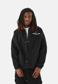 Chaqueta bomber negra de tejido suave, con cierre de botones a presión, puños elásticos y logo blanco "PROJET X PARIS" en el pecho.