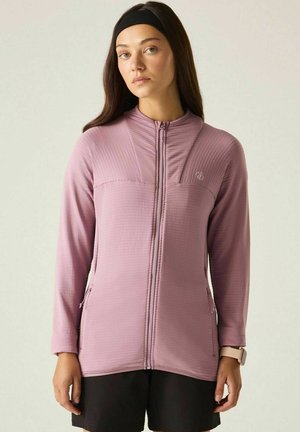 Mujer con cabello largo y oscuro lleva una chaqueta rosa con cremallera, pantalones cortos negros, una cinta negra para la cabeza y un reloj inteligente beige sobre un fondo liso claro.