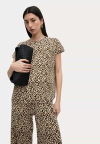 Top s leopardím vzorem a odpovídající kalhoty v béžové a černé barvě, vyrobené z texturované látky. Modelka drží černou tkanou kabelku.