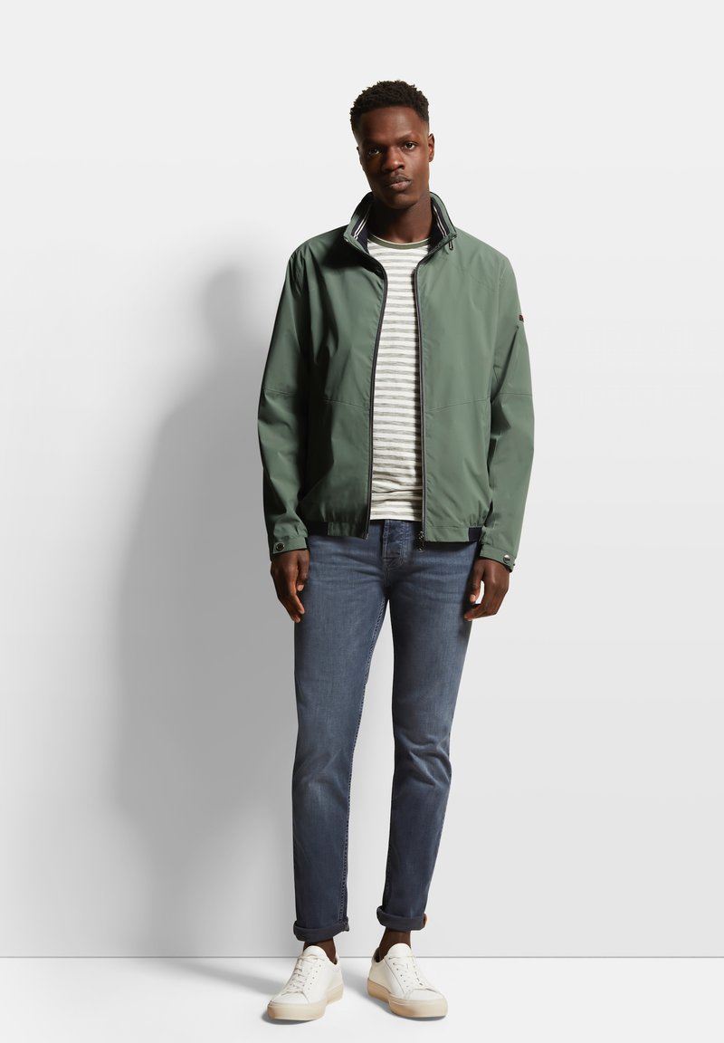 Veste légère verte avec un col haut, associée à une chemise rayée, un jean bleu et des baskets blanches. Texture lisse, design décontracté.