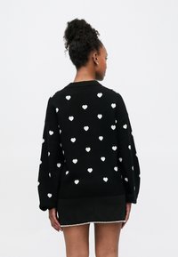 Maglione nero lavorato a maglia con motivi a cuore bianchi, caratterizzato da una vestibilità comoda e scollo a coste. La modella indossa una gonna mini nera con rifinitura di perle.