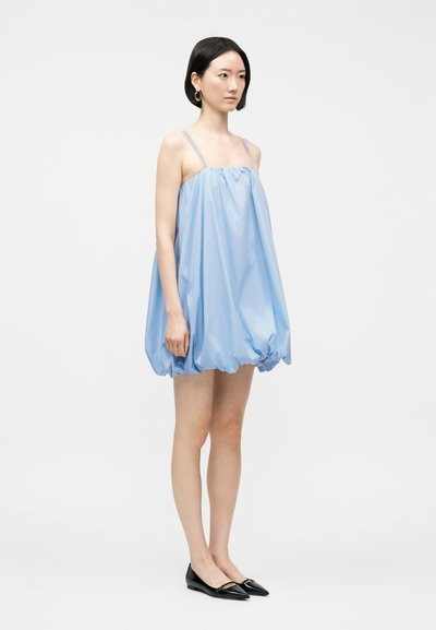 Femme debout vêtue d'une robe bulle ample bleu clair sans manches à fines bretelles, portant des ballerines noires pointues, sur un fond blanc.