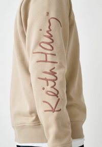 Beige sweatshirt med rund hals og ribstrikkede manchetter, med fremtrædende brun "Keith Haring" tekst på ærmet. Glat bomuldsstof.