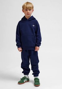 Marineblauer Hoodie mit Kängurutasche und weißem Logo, kombiniert mit passenden Jogginghosen mit elastischen Bündchen, getragen mit grünen Sneakers.