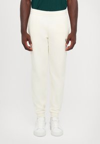 Cream-colored sweatpants made of soft fabric, featuring an elastic waistband and ribbed cuffs, with a small logo on the left side.