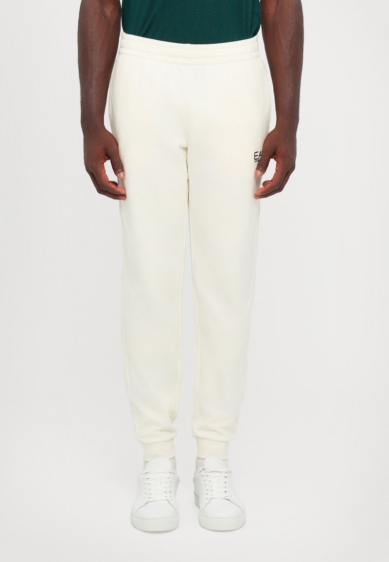 Cream-colored sweatpants made of soft fabric, featuring an elastic waistband and ribbed cuffs, with a small logo on the left side.
