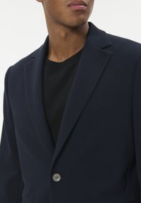 Tengerészeti struktúrával rendelkező blézer, egyetlen ezüst gombos záródással, lefelé hajlított kowernyakkal és alatta fekete kereknyakú pólóval.