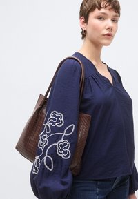 Μπλούζα ναυτικού μπλε με μακριά, φουσκωτά μανίκια που έχουν λευκή κεντητή ανθοδέσμη. Το μοντέλο κρατά μια καφέ τσάντα με υφή.