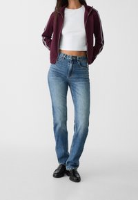 Burgundy zip-up hoodie med vita ränder, vit croptopp, ljusblå högmidjade jeans och svarta loafers. Slät textur, avslappnad passform.