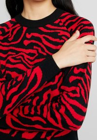 Černý svetr s červeným zebrovým pruhovaným vzorem, pružnými manžetami a klasickým kulatým výstřihem, který zvýrazňuje jemnou pletenou strukturu.