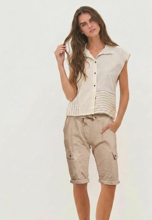 Femme aux longs cheveux bruns portant une chemise beige sans manches à boutons et un short cargo beige longueur genou, debout avec une main dans la poche.