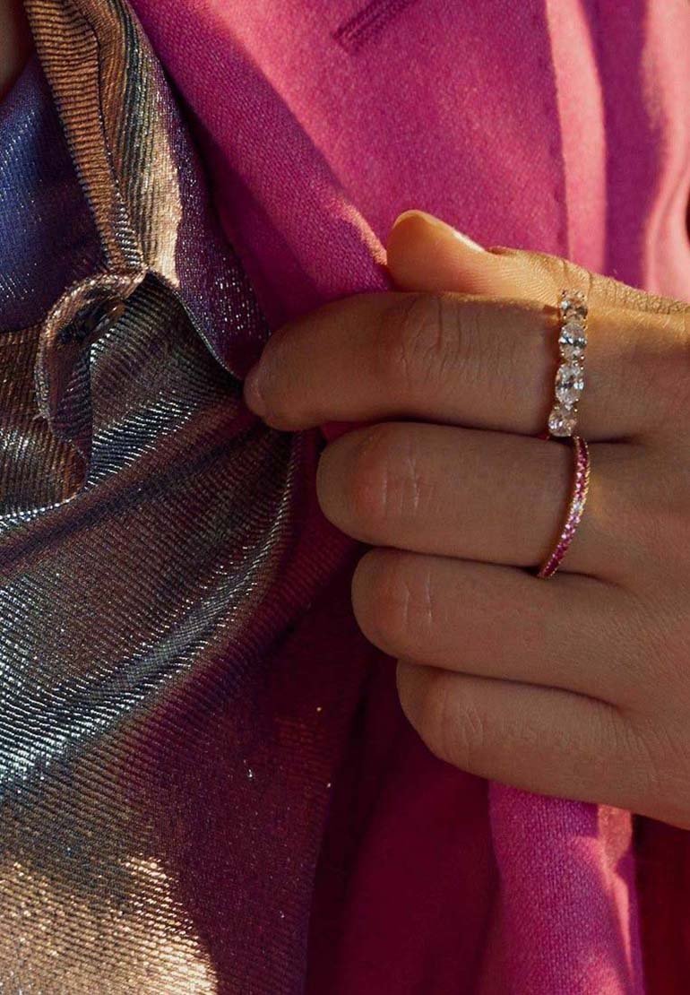 Silberner Ring mit drei klaren Steinen und einem pinken Band; Hand hält glattes, strukturiertes Silber und lila Stoff vor einem strahlend pinken Hintergrund.