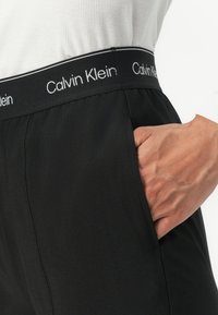 Čierne športové nohavice s elastickým pásom a bielym nápisom "Calvin Klein". Viditeľná bočná vrecko, hladká textúra po celom povrchu.