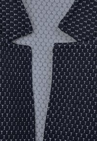 Námorně modrá texturovaná pletenina s bílým vzorem, s otevřeným límcem a štěpeným designem. Pozadí tvoří světlejší, síťovinový materiál.