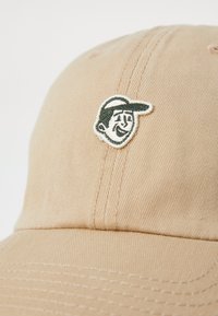 Beige Baumwollkappe mit strukturiertem Stoff, gebogenem Schirm und einem gestickten Cartoon-Gesicht-Logo in dunkelgrün und weiß.