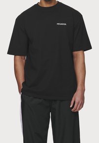 Černé bavlněné tričko s krátkým rukávem a kulatým výstřihem. Logo "PEGADOR" v bílé barvě na levé straně hrudi. Volný střih, hladká textura.