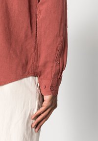 Camisa de manga larga color óxido hecha de tela texturizada, con puños abotonados y detalle de apertura lateral, acompañada de pantalones de color claro.
